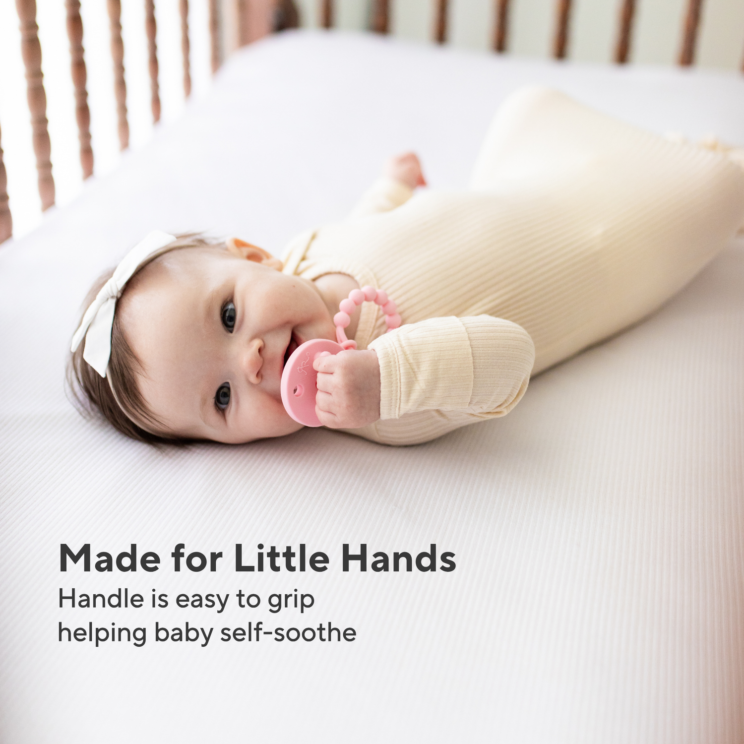Baby lying on a crib with a pink teething toy, wearing a white outfit.