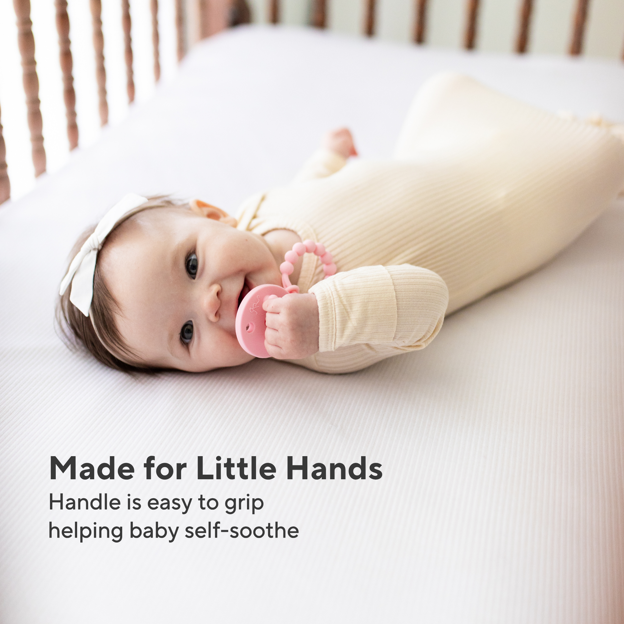Baby lying on a crib with a pink teething toy, wearing a white outfit.