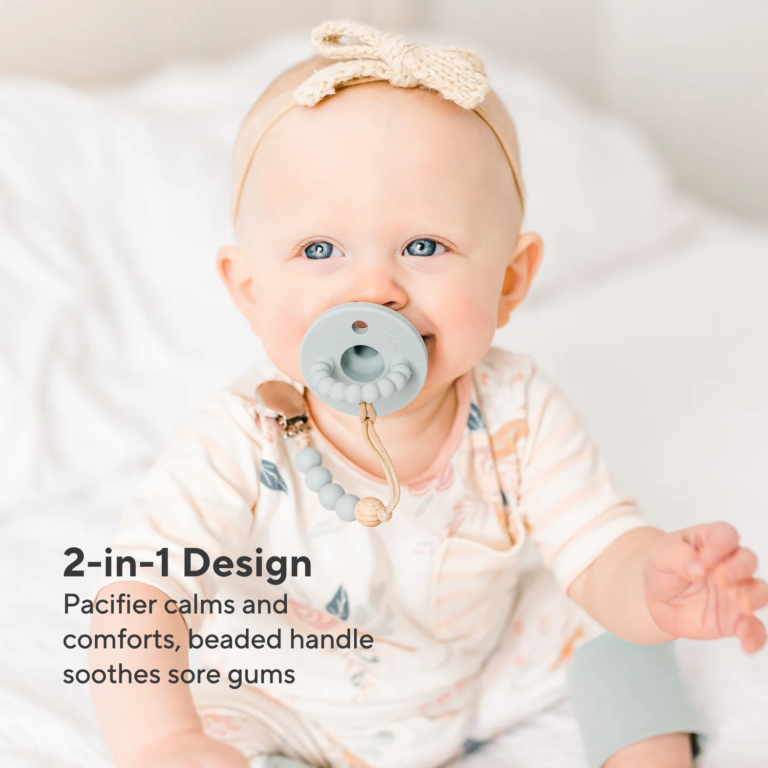 Baby wearing a pacifier with a beaded handle, sitting on a white blanket.