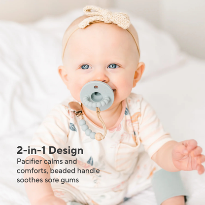 Baby wearing a pacifier with a beaded handle, sitting on a white blanket.