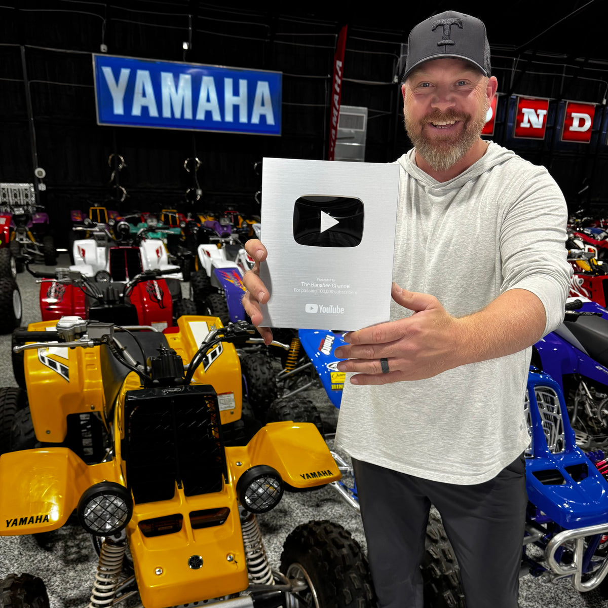 A smiling man in a gray hoodie and cap holds up a large silver YouTube Play Button award plaque in front of a busy indoor show floor with Yamaha and ATV products in the background.