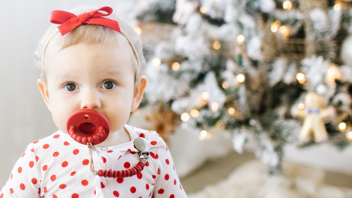 A baby wearing a red bow headband and a white onesie with red polka dots, holding a red pacifier in their mouth, with a blurred Christmas tree in the background.