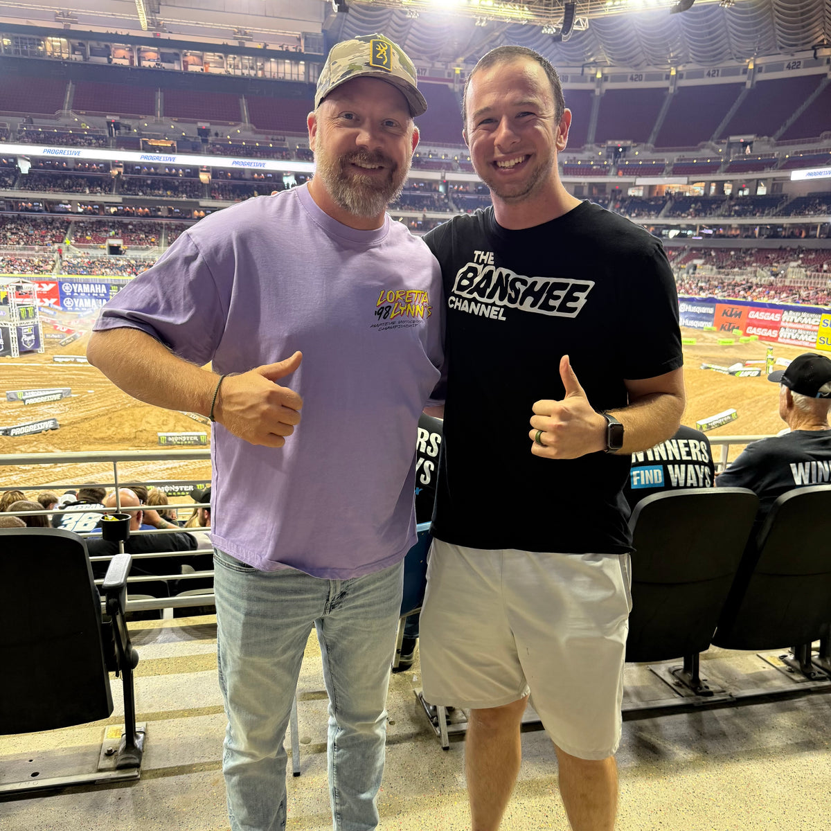 Two men posing together in an arena setting, with one wearing a lavender t-shirt and jeans and the other wearing a black &