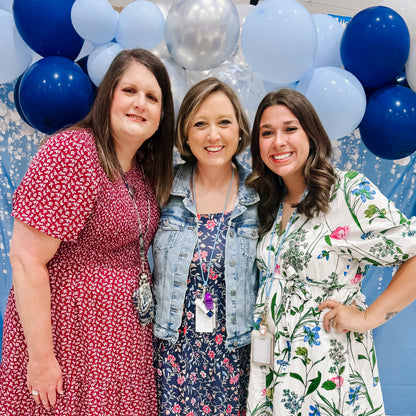 Three women posing together with blue, silver, and white balloons in the background.