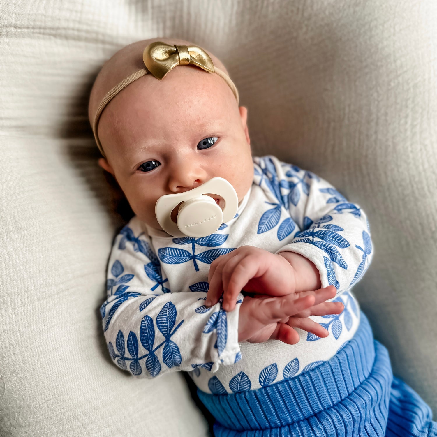 A baby wearing a blue and white patterned long-sleeve top, a blue ribbed knit bottom, a beige headband with a gold bow, and a pacifier, lying on a textured cream fabric.