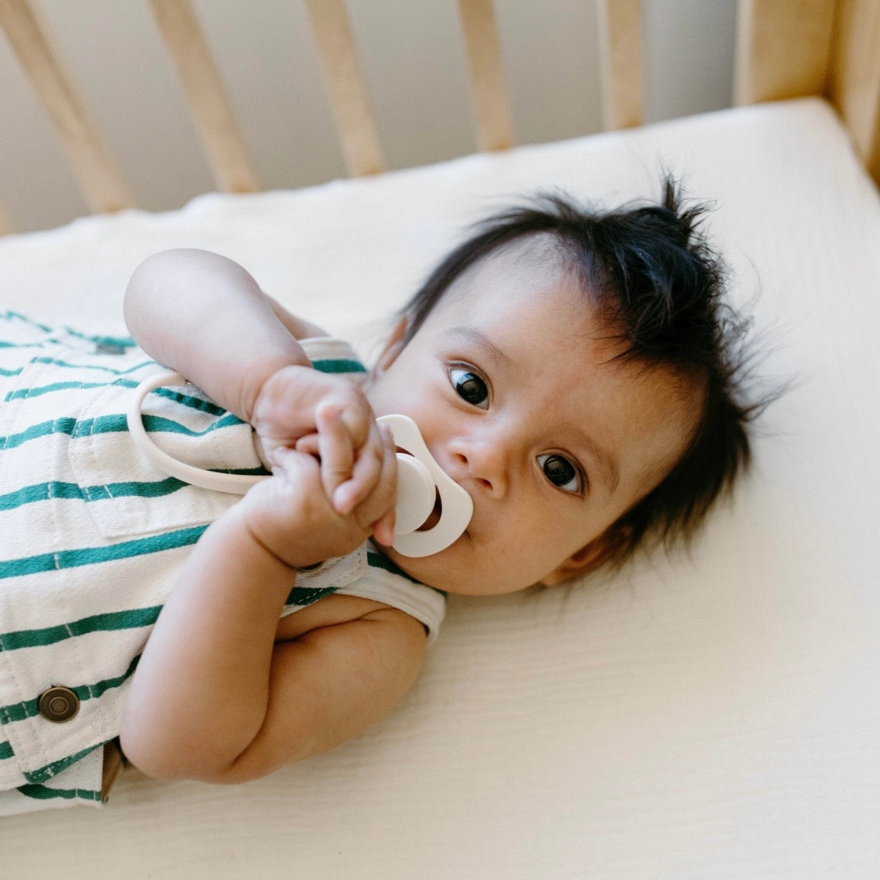 A baby lying on a cream mattress in a wooden crib, wearing a green and white striped romper and sucking a white pacifier.