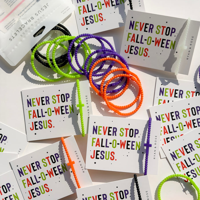 A styled flat-lay of colorful braided hair ties and matching printed cards on a white background. The hair ties come in black, neon green, orange, and purple, and the cards feature the phrase 'NEVER STOP FALL-O'WEEN JESUS.