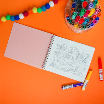 A spiral-bound sketchbook with black line drawings on white paper, laid on a bright orange background with colorful markers, a cup of markers, and a string of pom-poms.