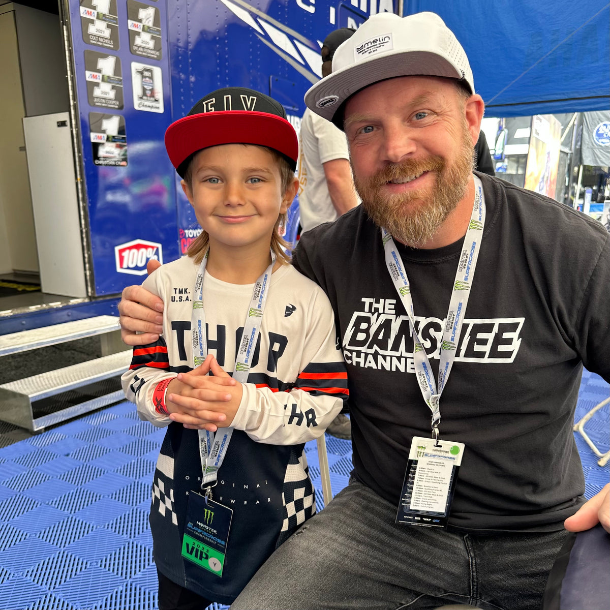 A man and a young boy posing together in front of a blue race truck at a motorsport event. The boy is wearing a red and black cap, a white and navy Thror long-sleeve jersey, and a lanyard, while the man wears a black &