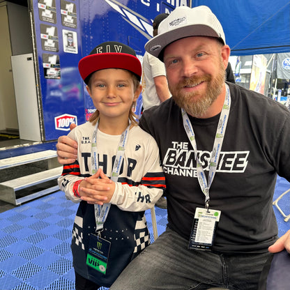 A man and a young boy posing together in front of a blue race truck at a motorsport event. The boy is wearing a red and black cap, a white and navy Thror long-sleeve jersey, and a lanyard, while the man wears a black &