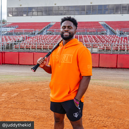 Man in orange hoodie holding a baseball bat on a baseball field with red seats in the background