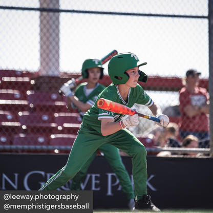 Child in green baseball uniform holding a bat on a field with spectators in the background.