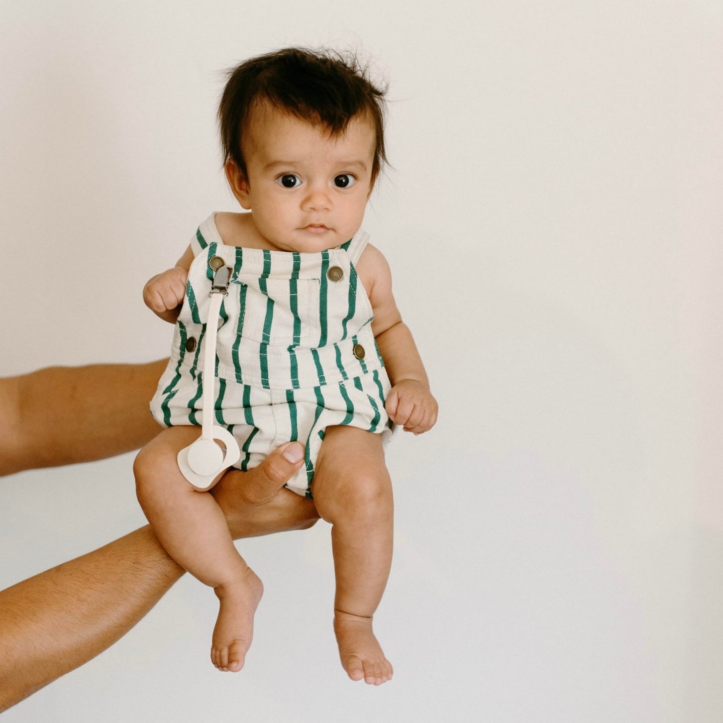 A baby wearing a green and white striped sleeveless romper is held up by an adult&