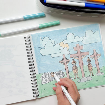 A hand holds a white marker over an open spiral notebook showing a colorful drawing of crosses, people, and clouds. Two markers lie on the white surface above the notebook, with a pen case partially visible at the top.