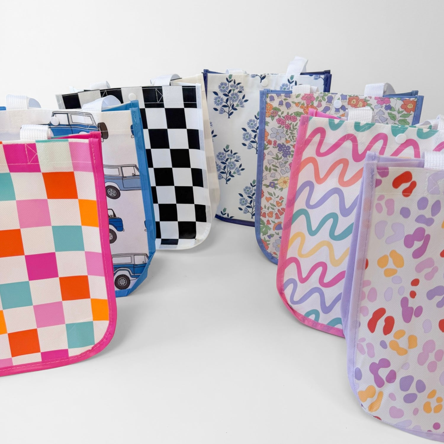A group of reusable fabric shopping bags with various colorful patterns, including checkered, floral, wavy, and animal print designs, arranged on a light gray studio background.