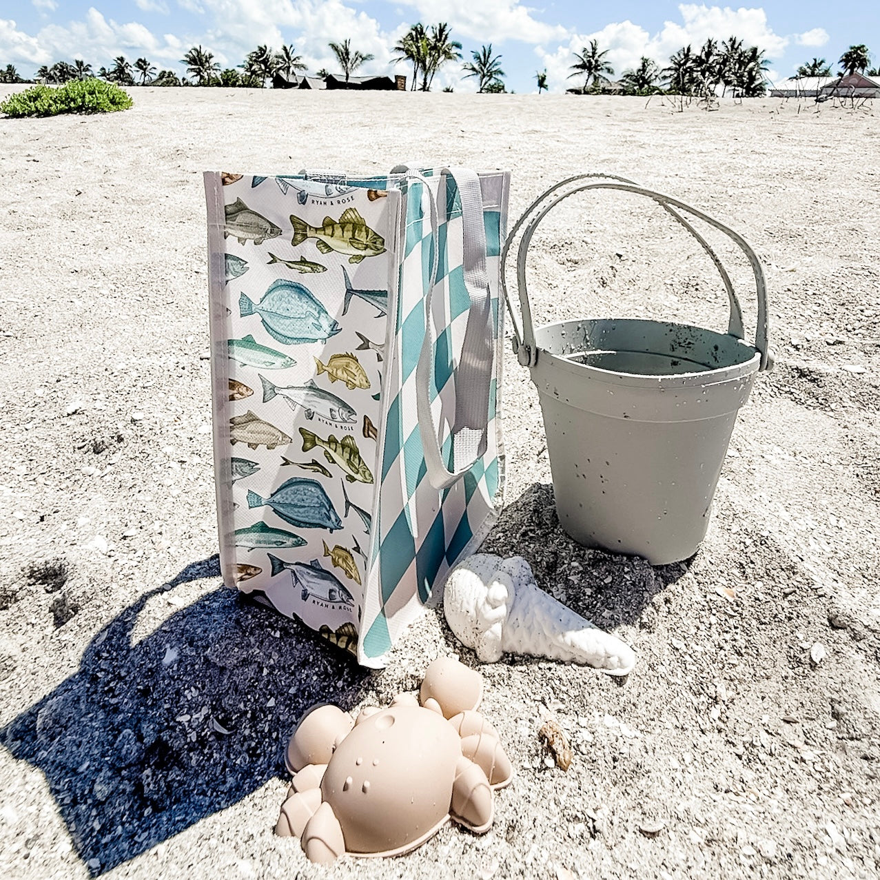 Beach scene with a bag, bucket, and sand toys on sandy ground with palm trees in the background.