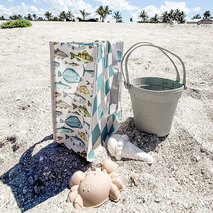 Beach scene with a bag, bucket, and sand toys on sandy ground with palm trees in the background.