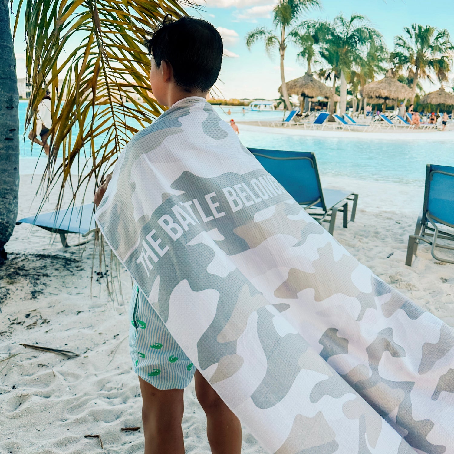 Person on a beach with a camouflage towel draped over them, featuring the text &