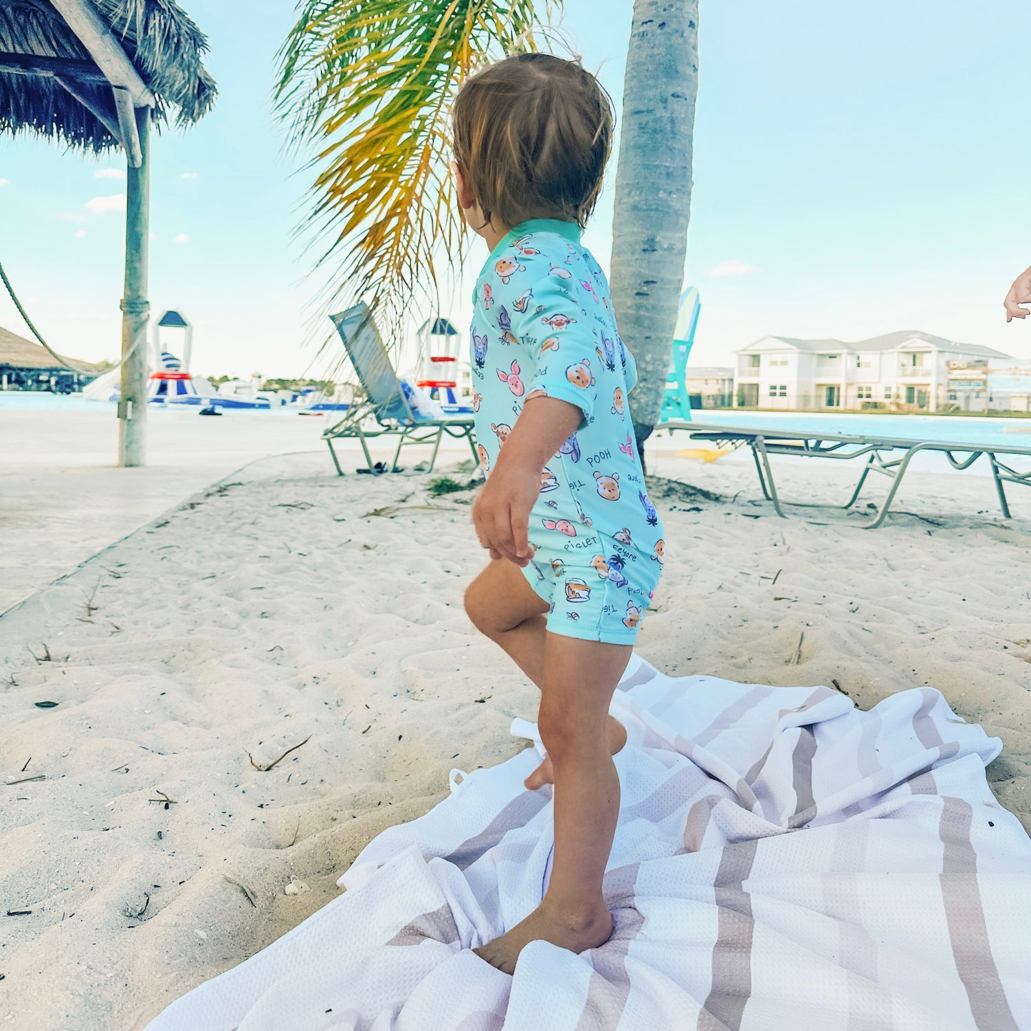 Child in a colorful swimsuit standing on a towel at the beach with palm trees and lounge chairs in the background.