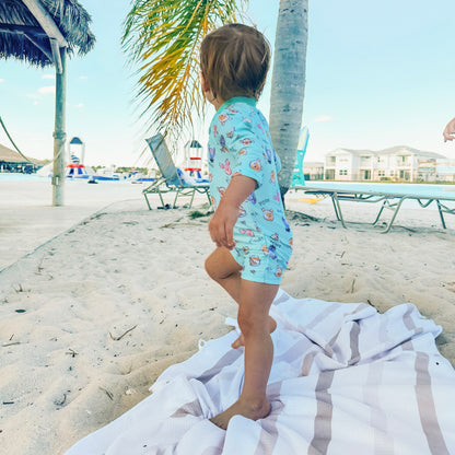 Child in a colorful swimsuit standing on a towel at the beach with palm trees and lounge chairs in the background.