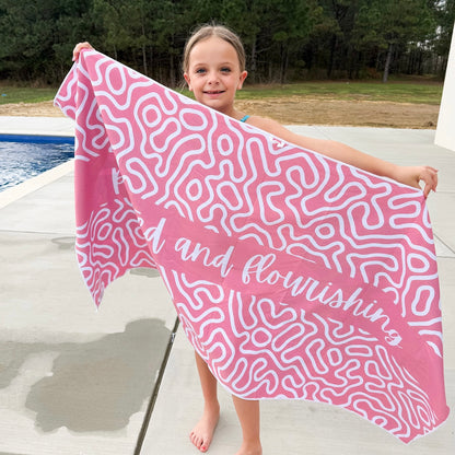Child holding a pink towel with white patterns and text by a poolside.