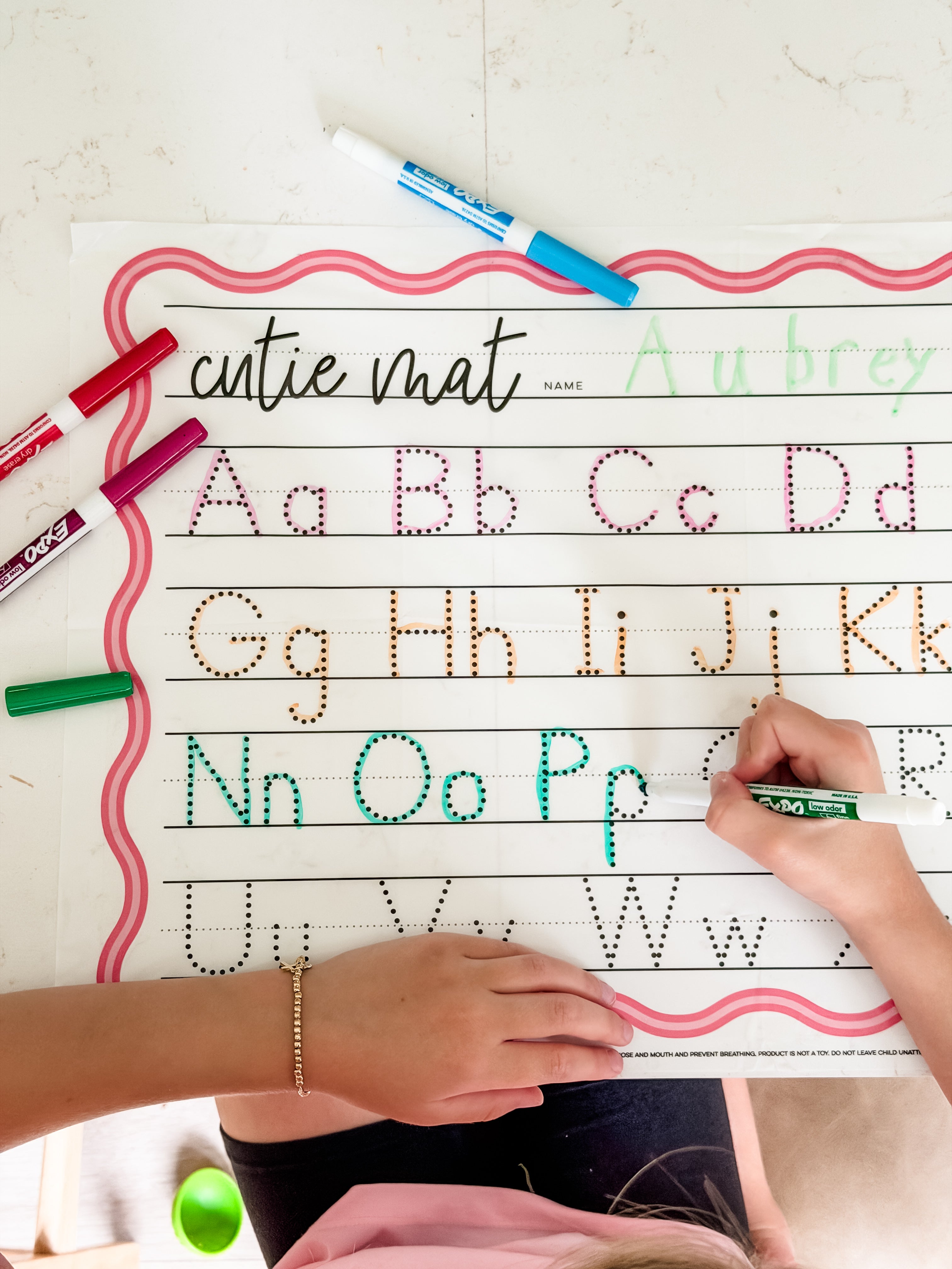Child writing on a cutie mat with alphabet tracing lines and colorful markers.