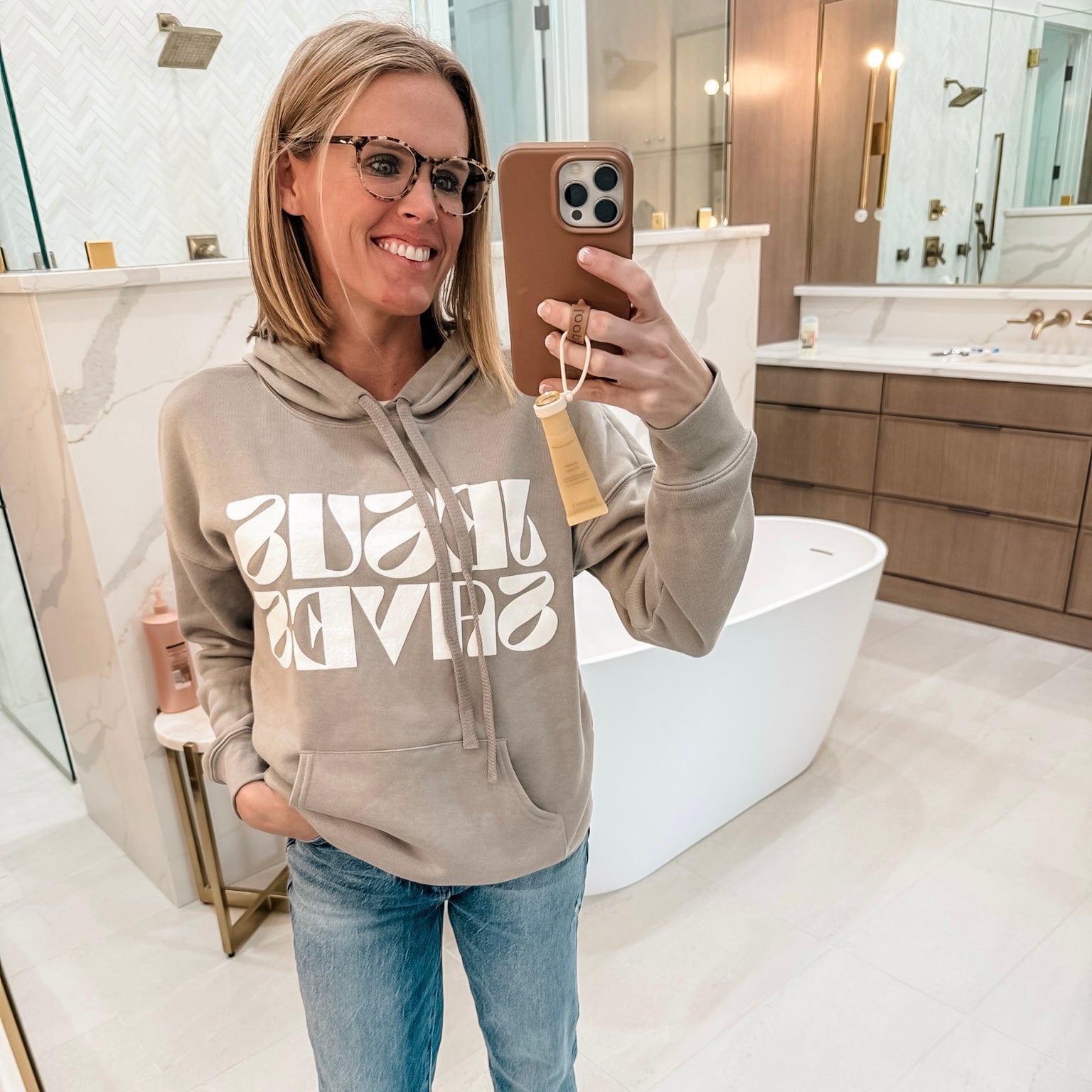 Woman taking a mirror selfie in a bathroom wearing a beige hoodie with text and blue jeans.