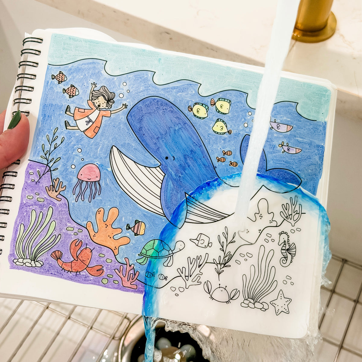 A hand holds a spiral-bound notebook showing a colorful underwater illustration, with water running over it to demonstrate water resistance. The notebook is placed over a tiled sink.