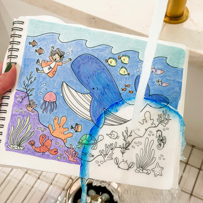 A hand holds a spiral-bound notebook showing a colorful underwater illustration, with water running over it to demonstrate water resistance. The notebook is placed over a tiled sink.