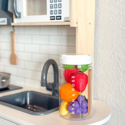 A clear plastic fruit storage jar with a white lid sits on a kitchen counter, filled with colorful toy fruits. The jar has the vertical text &
