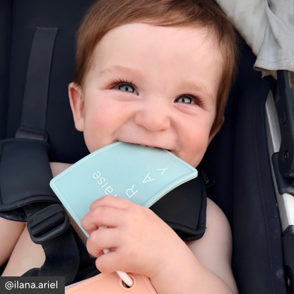 A baby wearing a black stroller seat belt and holding a light blue silicone shield with the brand name &