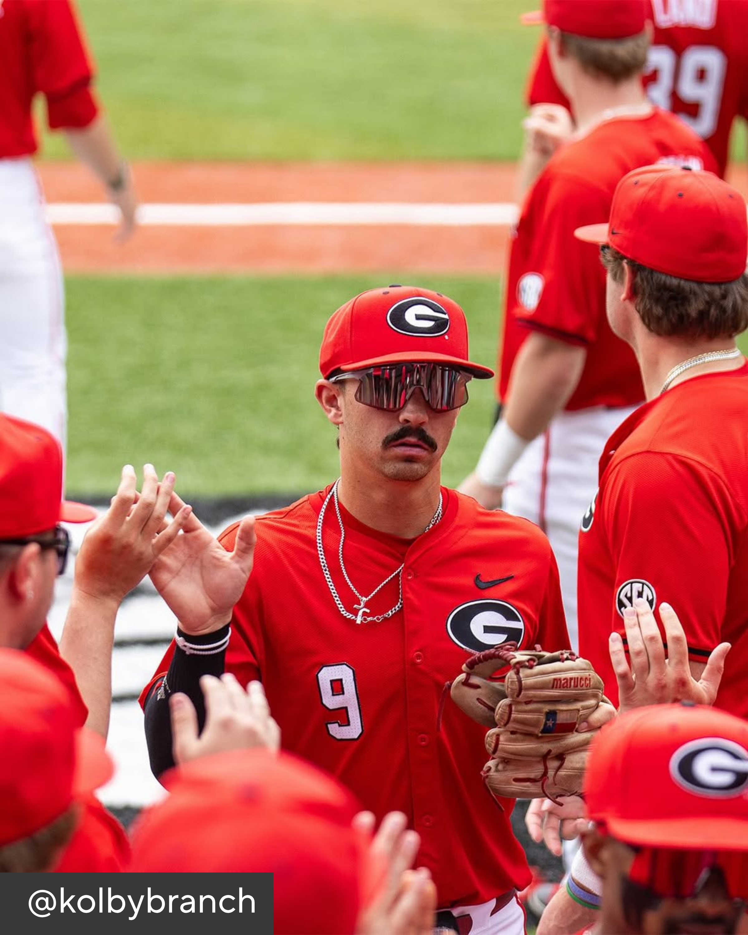 Baseball player in red uniform with sunglasses and mustache, surrounded by teammates on a field.