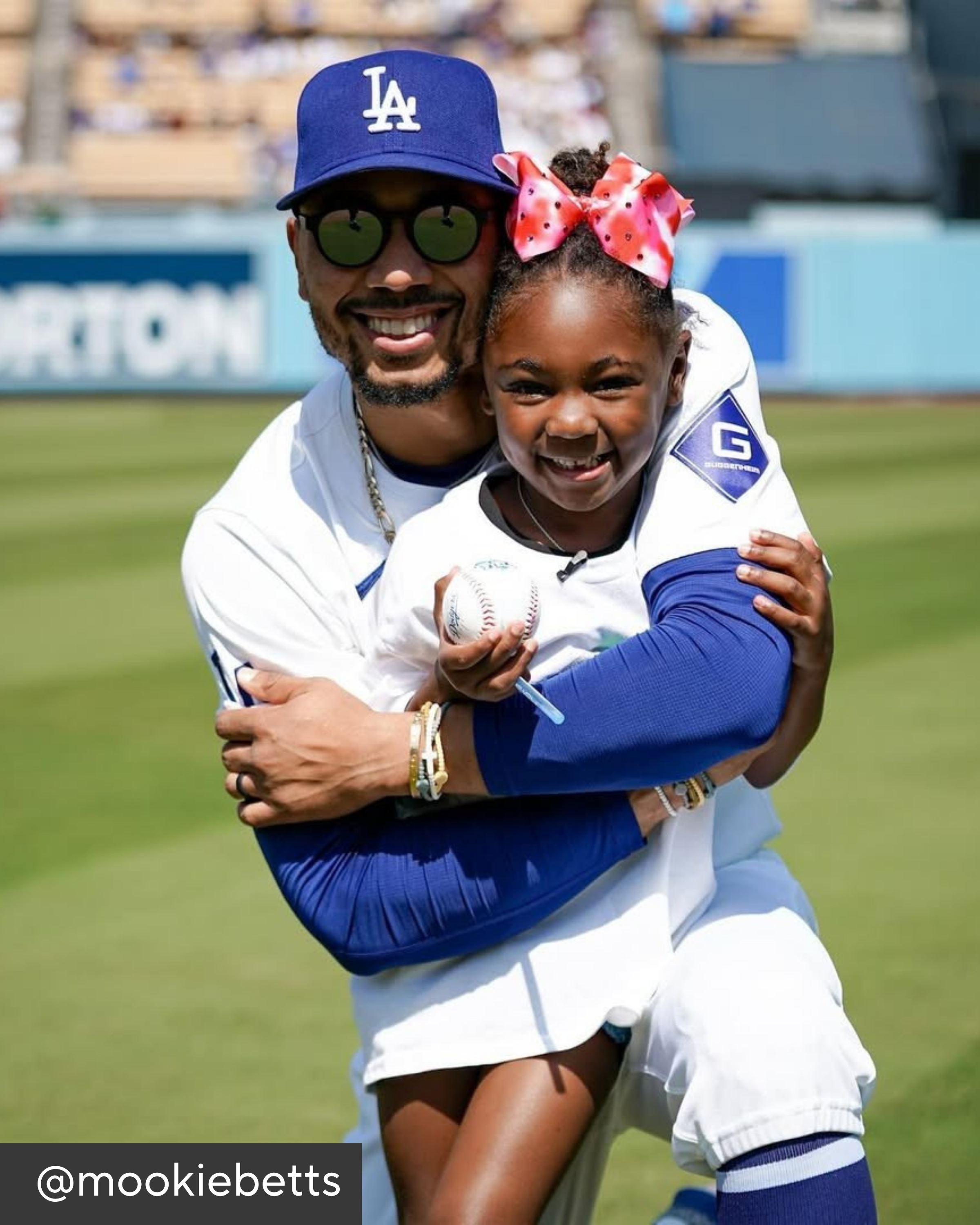Man and young girl hugging on a baseball field, both wearing sports jerseys.