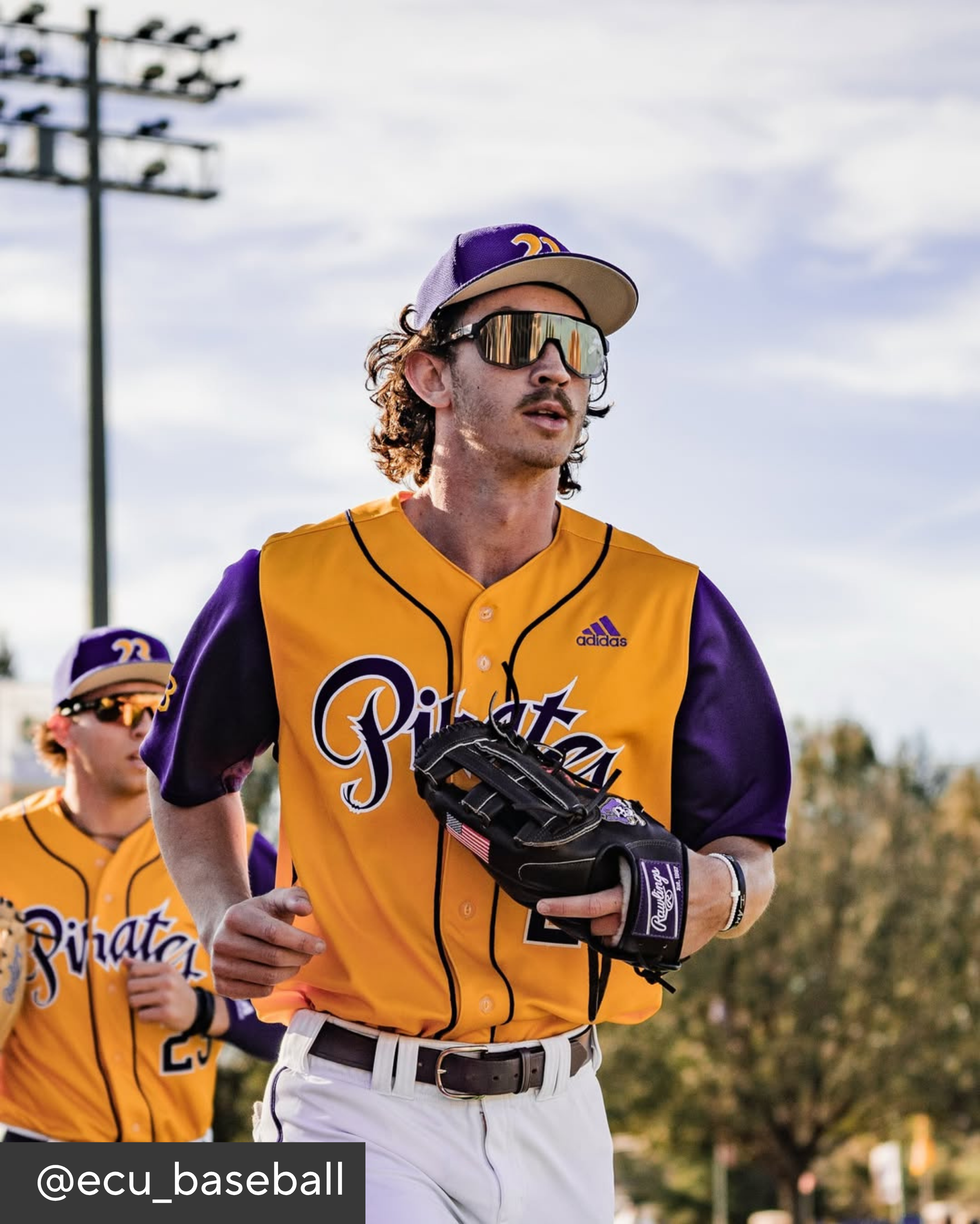 Baseball player in a yellow and purple uniform with sunglasses and a glove, standing outdoors.