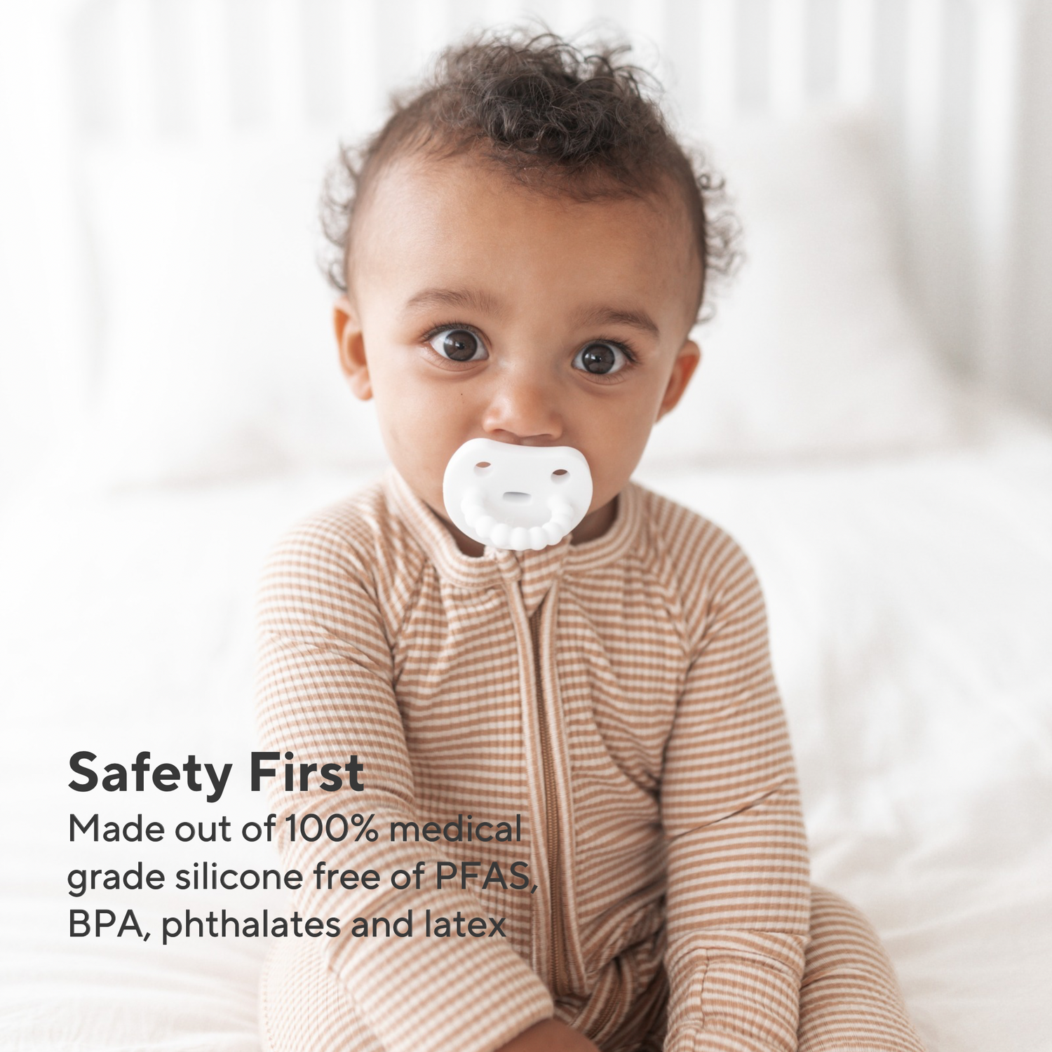 A baby wearing a beige and white striped zip-up onesie sits on a white bed, with a white silicone pacifier in their mouth. Text on the left reads, &