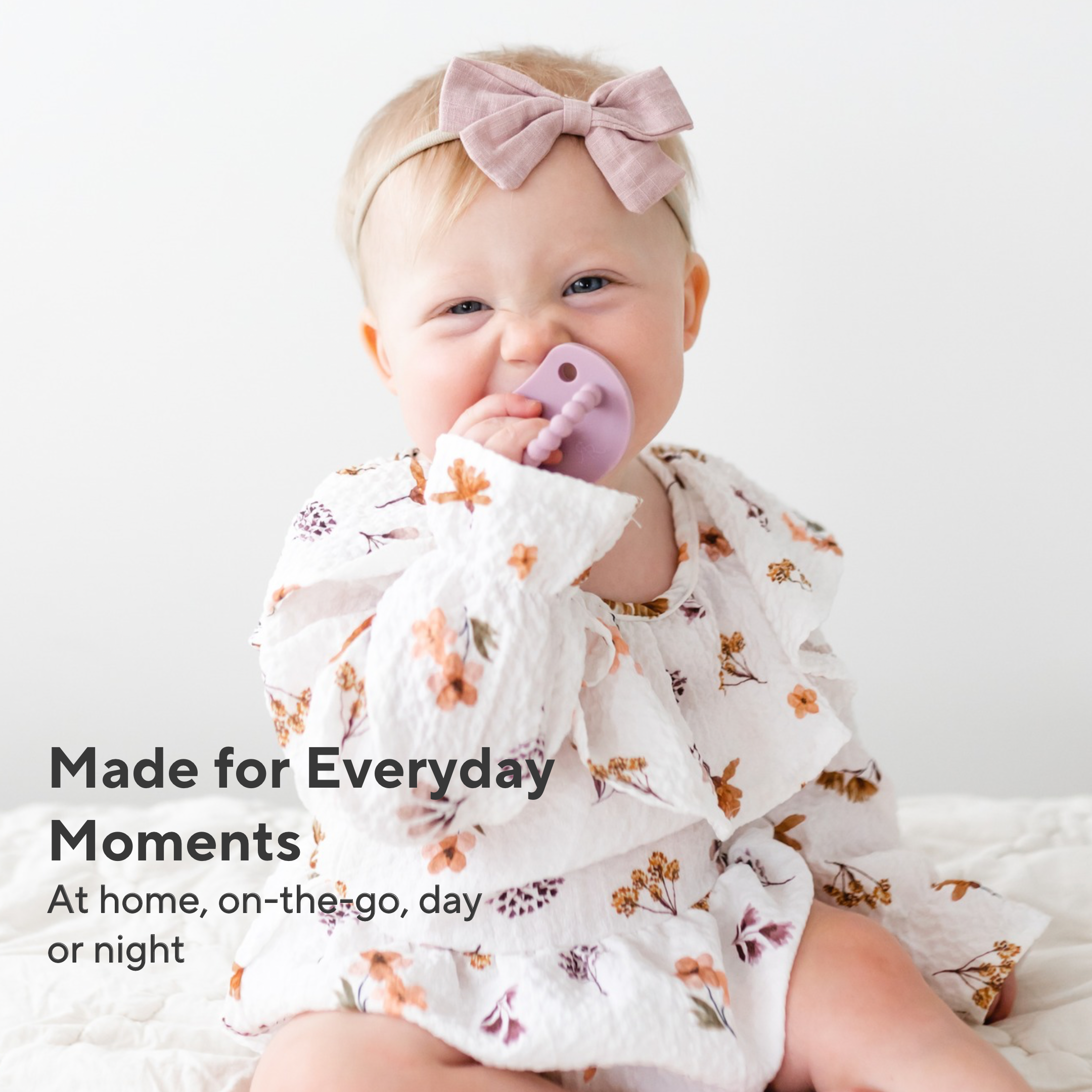 A baby wearing a white long-sleeve ruffled outfit with a floral print and a matching pink bow headband, sitting against a white background. The baby is holding a purple pacifier.