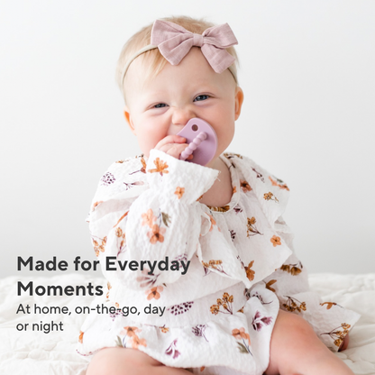 A baby wearing a white long-sleeve ruffled outfit with a floral print and a matching pink bow headband, sitting against a white background. The baby is holding a purple pacifier.
