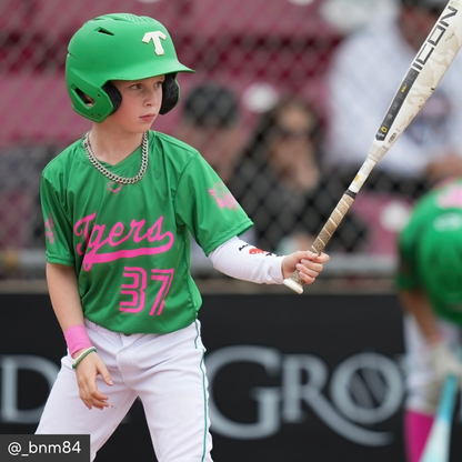 Child in green baseball uniform with pink lettering holding a bat, wearing a helmet.