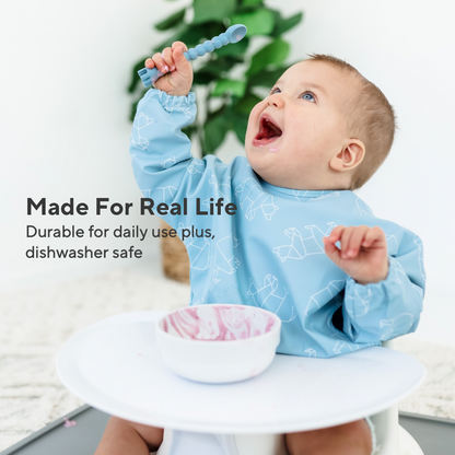 Baby in a blue bib sitting in a high chair with a bowl and spoon, surrounded by greenery.