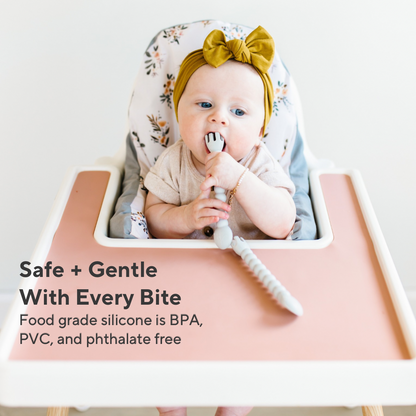 Baby in a high chair with a silicone spoon, wearing a floral headband, on a white background.