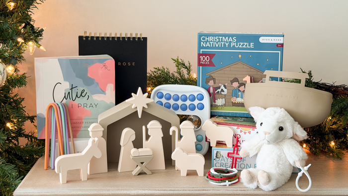 A styled holiday gift set on a tabletop featuring a wooden nativity scene, a plush white lamb toy, a boxed Christmas nativity puzzle, a notebook with the words 'Cutie, PRAY', colorful hanging ribbons, and other Christmas-themed items, with a decorated Christmas tree and warm string lights in the background.
