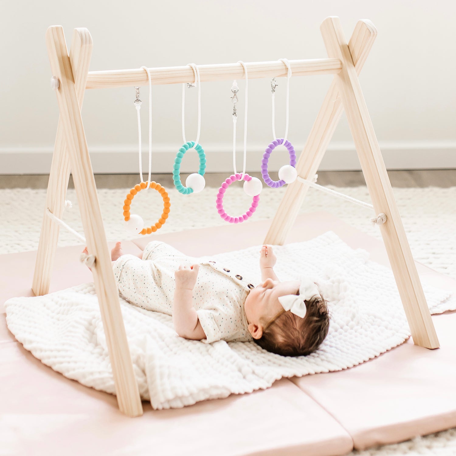 Baby laying underneath a play gym with Cutie Clasps hanging down attached to Cutie Teether Rattles.